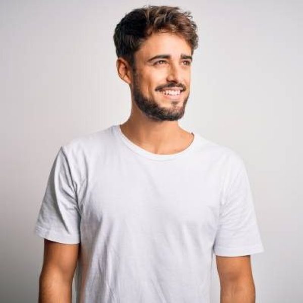 Young handsome man with beard wearing casual t-shirt standing over white background looking away to side with smile on face, natural expression. Laughing confident.